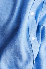 Camisa celeste de tejido liviano en mezcla de lino y viscosa, con cuello clásico, botones al frente, canesú de doble capa con pliegue de caja en la espalda, mangas largas con puños abotonados, bolsillo superior abierto y bajo redondeado.