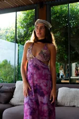 Vestido largo con estampado tie-dye en tonos violeta y fucsia, escote en V con detalles bordados dorados, cut-outs laterales bajo el busto y tirantes finos que se cruzan y atan en la espalda.