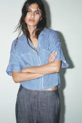 Camisa cropped de viscosa y lino a rayas verticales azules y blancas. Tiene cuello solapa, escote pico, manga corta con vuelta y bolsillos de plastrón.