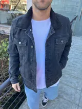 Chaqueta de jean negra con cuello de corderito negro, cierre frontal con botones metálicos, bolsillos con solapa abotonada en el pecho y bolsillos laterales.
