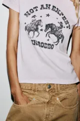 Camiseta color gris claro con cuello redondo, mangas cortas y hombros caídos. Estampado frontal con dos jinetes a caballo y la frase "Not an expert in rodeo".