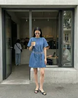 Vestido corto de denim azul, con cuello redondo y mangas cortas amplias. Presenta un corte ligeramente entallado y un cinturón de cadena metálica con eslabones circulares.