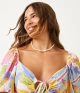 Top cropped de tejido tipo laise con estampado floral pixelado en tonos pastel (rosa, amarillo, azul, blanco). Presenta escote corazón con abertura central y lazo ajustable, y mangas cortas abullonadas.