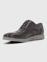 Zapato tipo Oxford o Brogue de cuero marrón oscuro, con puntera y laterales decorados con perforaciones (broguing). Posee cordones grises y una suela deportiva gruesa de goma color gris claro.