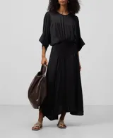 Vestido largo negro de mujer con manga japonesa, cuello caja en delantero y escote en espalda con nudo. Ajuste en cadera con godets y bajo asimétrico.