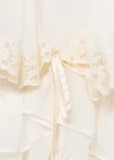 Blusa color crema con cuello en V, volados y detalles de encaje. Tiene mangas largas con puños elásticos y lazos.
