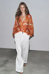 Camisa arrugada de ramio color naranja con estampado floral en tonos rosados y blancos. Tiene cuello redondo con escote pico ajustable con lazadas, mangas 3/4 y bajo asimétrico con aberturas laterales.