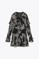 Vestido corto negro con estampado paisley en tonos beige y detalles dorados. Escote en V con lazos ajustables, mangas largas con puños elásticos y volantes en el mismo tejido en el escote y el ruedo.