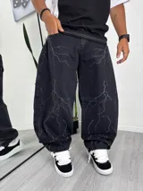Pantalón de jean negro, corte baggy o ancho, con lavado que simula un estampado de rayos o grietas en color gris claro.