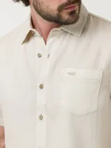 Camisa de manga corta color beige, con cuello clásico, cierre frontal con botones y bolsillo en el pecho izquierdo.