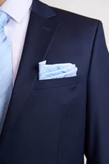 Traje de vestir azul marino, compuesto por saco de dos botones con solapa de muesca, dos bolsillos laterales con solapa y un bolsillo de ojal en el pecho, y pantalón de vestir con corte recto. Incluye camisa blanca, corbata celeste y pañuelo de bolsillo blanco con borde celeste.