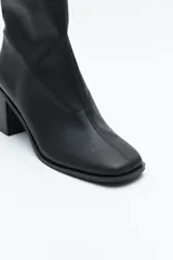 Bota corta de caña elástica, color negro con acabado charolado. Presenta puntera cuadrada y taco medio geométrico y ancho.