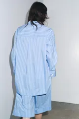 Camisa celeste con rayas verticales blancas, de corte amplio, cuello clásico, manga larga con puños y cierre frontal con botones.