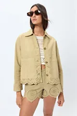 Sobrecamisa color beige claro, estilo chaqueta de trabajo o chore coat, con cuello camisero, botones frontales y dos bolsillos de parche en el pecho. Presenta detalles de bordado troquelado (broderie anglaise) en el ruedo inferior.