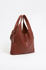 Bolso de mano color marrón de cuero con estructura blanda. Incluye asa extraíble, bolsillo interior y etiqueta de tejido. Presenta logo metálico BIMBA Y LOLA con acabados metálicos plateados.