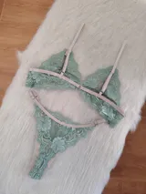 Conjunto de lencería de encaje floral color verde menta, con detalles y tiras ajustables en tono beige/tostado. Incluye un corpiño triangular sin aro y una bombacha tipo colaless o tanga.