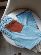 Pantalón de jean celeste con cintura elastizada, corte recto, bolsillos laterales y aplique de cuero marrón en el frente. Presenta botones metálicos en los laterales y etiqueta de cuero en la parte posterior.