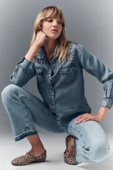 Camisa de denim celeste con apliques de piedras plateadas en los hombros, corte recto, cuello camisero, botones metálicos y bordado Rapsodia en la espalda.