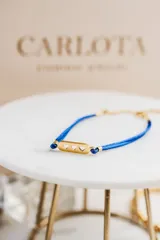 Pulsera con hilo azul brillante y detalles dorados en acero quirúrgico. Dije rectangular dorado con tres corazones calados.