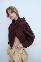 Trench corto color negro, con cuello solapa y capucha desmontable mediante botones. Manga larga tipo murciélago acabada en trabilla ajustable. Bolsillos laterales. Forro interior combinado. Cierre frontal con botones. Tejido repelente al agua.