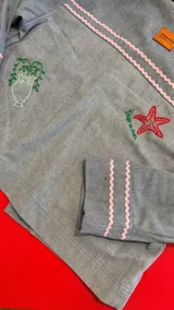Chaqueta de denim celeste con bordados artesanales. Presenta bordados de flores rojas y verdes en el frente, un delfín/sirena en el bolsillo derecho y detalles de ondas en color rosa pálido en el cuello, hombros y puños. Los bordes de los bolsillos tienen flecos dorados.