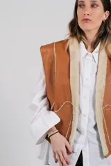 Chaleco reversible de piel de cordero color beige claro, con forro de cuero marrón visible en el borde. Se lleva sobre una camisa blanca de manga larga.