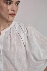 Camisa blanca con estampado de cruces, cuello mao y mangas 3/4 con detalle de pliegues en los hombros. Cierre frontal con botones.
