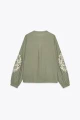 Blusa verde oliva de algodón con cuello redondo y escote en pico con cordones y borlas. Tiene manga larga con bordados florales en los hombros, jaretas y entredoses delanteros. Cierre frontal con botones.