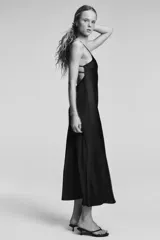 Vestido midi negro de tejido satinado, con escote en pico y tirantes finos ajustables. Presenta un detalle de tiras cruzadas en la espalda descubierta y cierre con cremallera metálica.