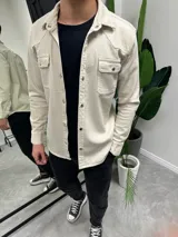 Chaqueta color beige de corte recto, con cuello camisero, cierre frontal con botones metálicos a presión, dos bolsillos con solapa abotonada en el pecho y mangas largas con puños abotonados.