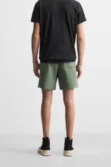 Bermuda color verde militar de corte relajado, confeccionada en tejido de algodón. Tiene cintura elástica ajustable con cordón interior, bolsillos frontales y bolsillo trasero de parche.