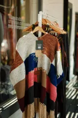 Ruana tejida tipo patchwork confeccionada en lana con diseño de bloques de colores en blanco, marrón, azul y rosado. Presenta un bordado central de sol, un parche de cuero con la inscripción 'José Ignacio' y terminación con flecos en el ruedo.