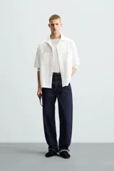 Sobrecamisa negra de tejido con elasticidad, cuello solapa y manga corta. Tiene dos bolsillos de plastrón en el pecho y cierre frontal con botones a presión.