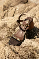 Sandalia de cuero color marrón con taco bloque medio y punta cuadrada. Presenta tachas esféricas doradas en las tiras y hebilla de ajuste en el tobillo.