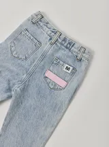 Pantalón de jean celeste con efecto lavado, corte recto y largo a la rodilla. Presenta una cinta vertical color violeta en la pierna izquierda, una cinta vertical color verde en la pierna derecha y una cinta horizontal color rosa en la parte inferior de ambas piernas.