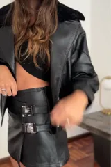 Minifalda negra de cuero sintético, tiro bajo, con corte irregular y detalle de dos hebillas plateadas en el lateral.