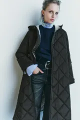Chaqueta larga acolchada color negro con capucha y manga larga. Tiene bolsillos delanteros y bajo asimétrico con aberturas laterales. El cierre frontal es con botones ocultos por solapa.
