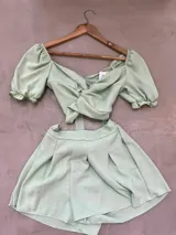 Conjunto color ocre compuesto por un top corto con escote en V, mangas cortas abullonadas y nudo en el frente, y un short de tiro alto con cintura elastizada. Incluye un cinturón blanco con tachas y hebilla dorada.