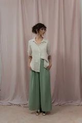 Camisa color crudo confeccionada en lino, de corte holgado, con cuello clásico, mangas cortas con dobladillo y cierre frontal con botones. Presenta detalle de pliegues verticales en la parte frontal.