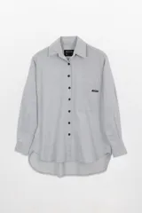 Camisa gris claro de tela Oxford, corte amplio, con cuello clásico, cierre frontal con botones negros, manga larga con puños abotonados, bolsillo de parche en el pecho y ruedo redondeado.