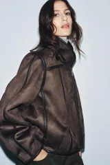 Cazadora de estilo aviador con cuello subido, manga larga y cierre frontal asimétrico con cremallera. Presenta un diseño de doble faz con exterior de efecto ante y forro interior de borreguillo a tono, con detalles de ribetes en cuero sintético.