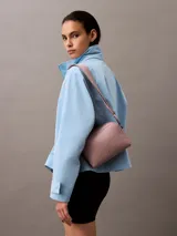 Bandolera rectangular pequeña de cuero sintético color lila con logo CK grabado al frente, cierre superior con doble cremallera y correa de tela ajustable con logo.