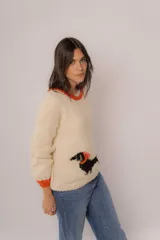 Sweater tejido color crema con cuello y puños acanalados en color naranja. Presenta un diseño bordado de un perro salchicha negro con un moño naranja.