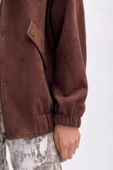 Chaqueta tipo bomber de antelina color marrón oscuro, con cuello mao, cierre frontal de botones a presión y puños y cintura elásticos. Posee bolsillos delanteros con solapa y botón a presión.