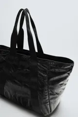 Bolso tipo shopper negro con acabado metalizado. Tiene un compartimento principal con cierre de cremallera y un bolsillo interior de malla con cremallera.