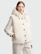 Chaleco Moncler modelo Arve en color blanco crema, con frente y capucha de pelo sintético. Presenta un diseño acolchado con cierre frontal de botones a presión negros, capucha ajustable con cordones y el logotipo de la marca bordado en el pecho.