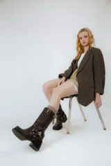 Bota de cuero color marrón oscuro, de caña media, con punta cuadrada y taco bloque. Presenta correas de cuero con hebillas metálicas plateadas alrededor del tobillo y en la parte superior de la caña.