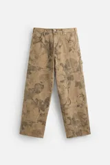 Pantalón de jean color beige con estampado abstracto floral en tonos marrones, de corte amplio, con bolsillos delanteros y traseros, y detalle de tira multifunción en la pierna. Cierre frontal con cremallera y botón.