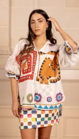 Camisa blanca con estampado de motivos étnicos en colores naranja, rojo, violeta, verde y azul. Tiene cuello camisero, mangas cortas con volados de encaje y cierre frontal con botones.