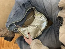 Cartera tipo shoulder bag con forma de banana, confeccionada en cuero metalizado color plateado. Tiene asa corta de cuero y asa larga desmontable de cadena metálica. El interior está forrado y cuenta con un bolsillo.